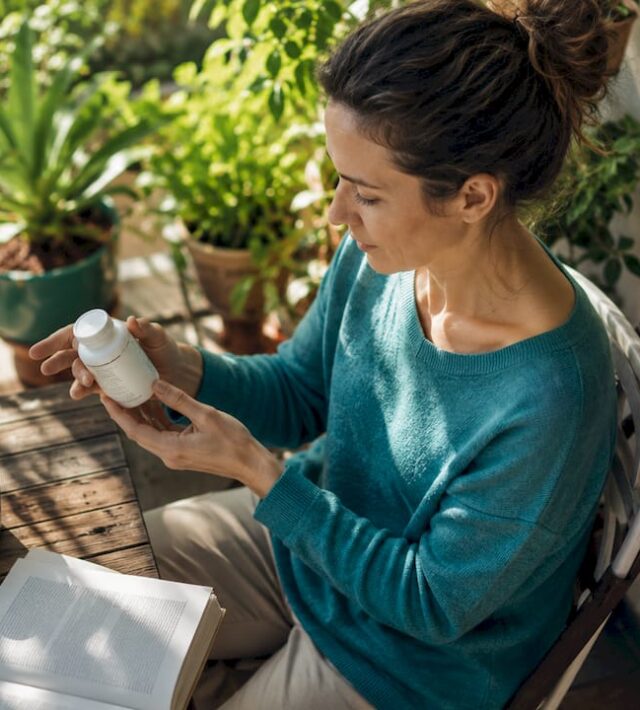 O femeie răsfoiește cu atenție eticheta unei sticle de vitamina D, într-un living plin de lumină naturală.