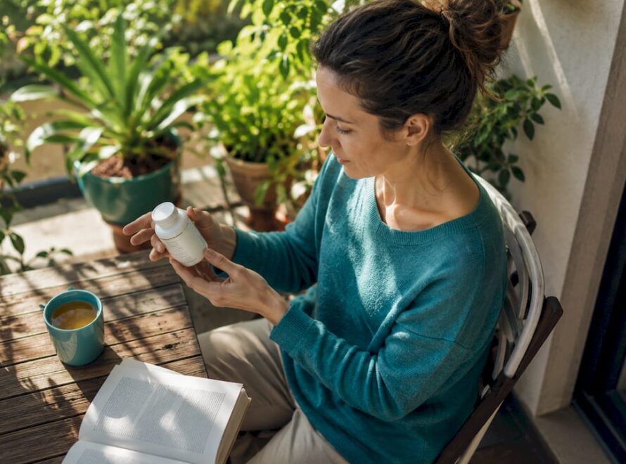 O femeie răsfoiește cu atenție eticheta unei sticle de vitamina D, într-un living plin de lumină naturală.