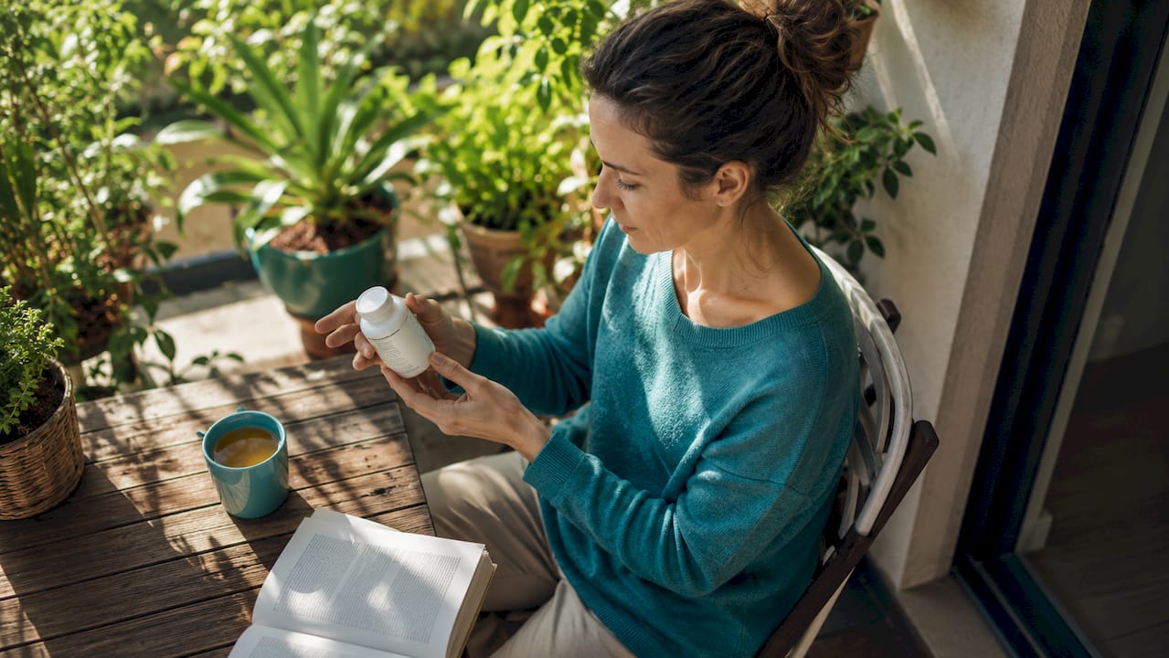 O femeie răsfoiește cu atenție eticheta unei sticle de vitamina D, într-un living plin de lumină naturală.